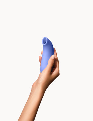 Hand holding a sleek, curved blue Dame Aer Air Pulse Stimulator with a soft silicone tip against a white background.