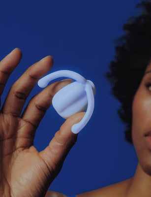 Hand holding a pale blue Dame Eva hands-free wearable vibrator against a solid blue background.
