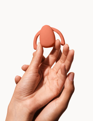 Hands holding a compact, coral-colored Dame Eva hands-free wearable vibrator against a white background.