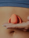 Close-up of a hand holding a coral-colored Dame Eva hands-free wearable vibrator against a bare abdomen.