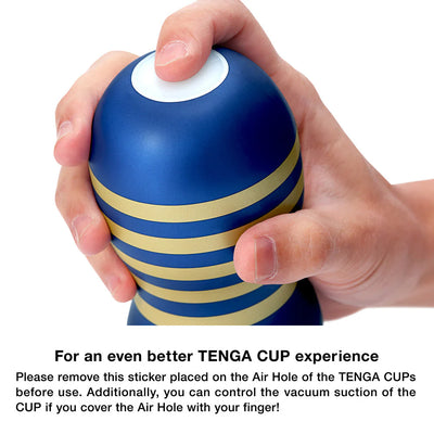 Hand holding a blue and gold striped Tenga Premium Original Vacuum Cup with a finger covering the air hole on top.
