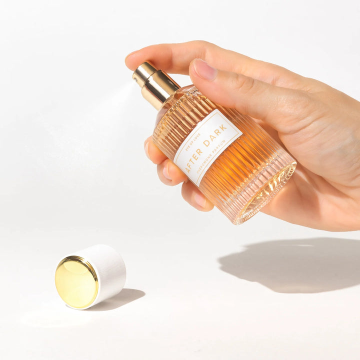 Hand spraying Eye of Love After Dark pheromone parfum in a ribbed glass bottle with gold-tone spray and white cap.