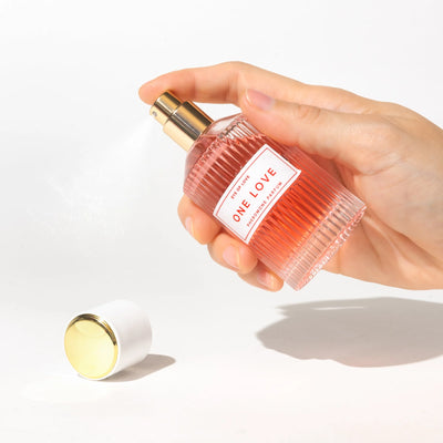 Hand holding and spraying Eye of Love One Love pheromone perfume bottle with gold and white cap on white background.