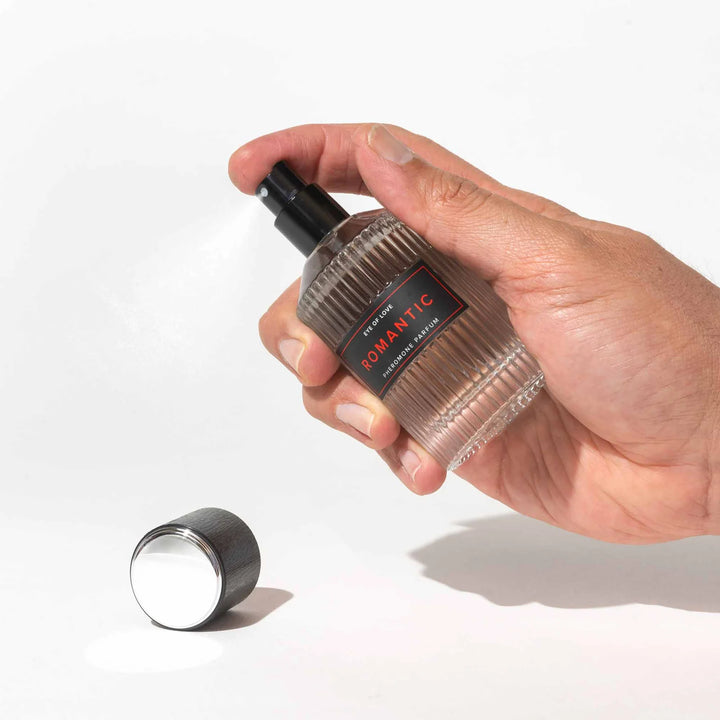 Hand holding and spraying Eye of Love Romantic pheromone perfume bottle with cap placed nearby on white background.