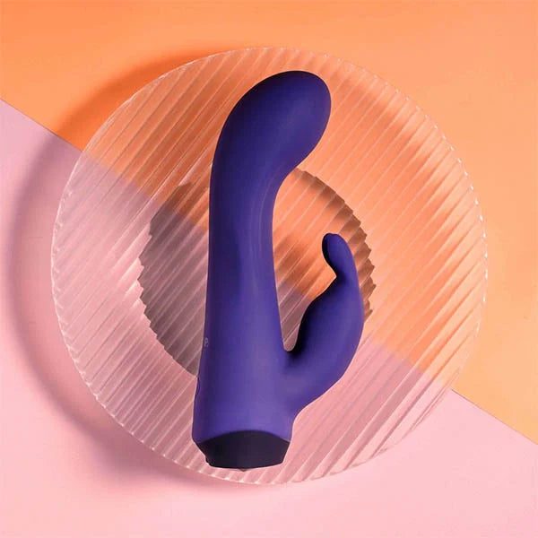Purple silicone dual stimulator vibrator with curved design on a textured clear plate against a peach and pink background.