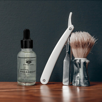 Eye of Love Natural Pheromone Beard Oil bottle with straight razor and shaving brush on wooden surface