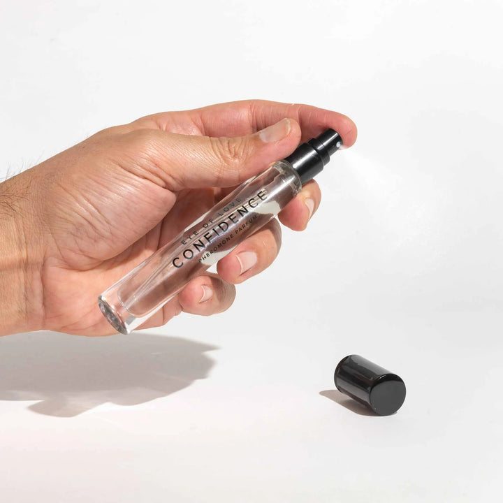 Hand holding Eye of Love Confidence pheromone perfume spray with black cap removed on white background.