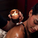 Hand holding lit Matchmaker massage candle near a woman's shoulder during a relaxing massage session.