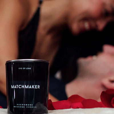 Eye of Love Matchmaker pheromone massage candle with red rose petals and a smiling couple in the background.