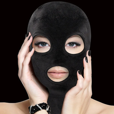 Close-up of a person wearing a black velvet mask with eye and mouth openings, hands gently touching the sides.