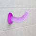 Glow-in-the-dark purple silicone pleasure toy with colorful confetti, curved shape, and suction cup base on tiled wall.