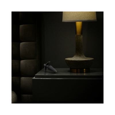 Black silicone vibrating plug placed on a dark bedside table next to a modern lamp in a dimly lit room.