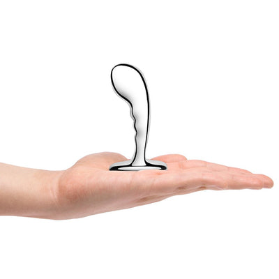 Hand holding a polished stainless steel prostate plug with a curved, textured design and wide base.
