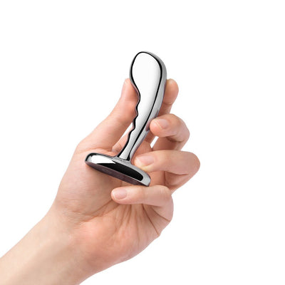 Hand holding a polished stainless steel prostate plug with a curved, ridged design and wide base.