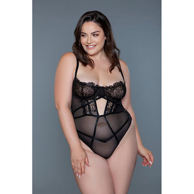 Plus size model wearing black lace and mesh BeWicked Cameryn bodysuit with underwire cups and adjustable straps against gray background.