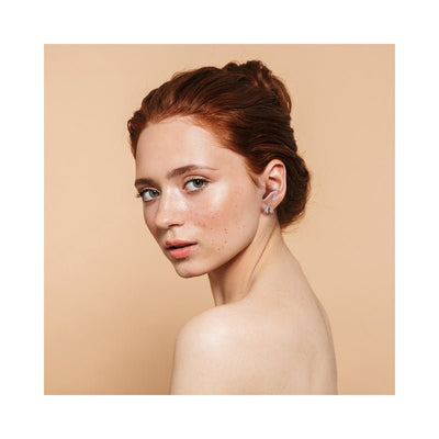 Close-up of a woman with red hair wearing delicate sterling silver Biird Clit Earrings against a beige background.