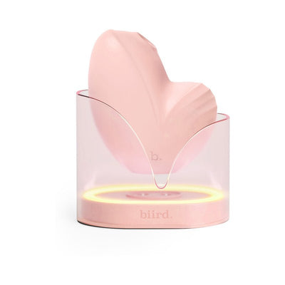Biird Namii clitoral stimulator in soft pink silicone resting in a translucent charging base with ambient light ring.