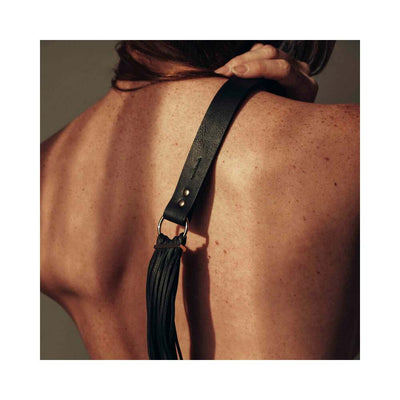 Close-up of a person holding a black vegan leather tassel flogger against their bare back.