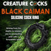 Black silicone cock ring shaped like a caiman with textured ridges and a grinning head on mossy green background.