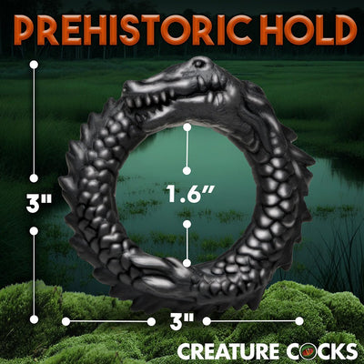 Black silicone cock ring shaped like a grinning caiman with textured ridges, shown with size measurements on a swamp background.