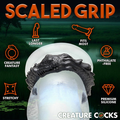 Black silicone cock ring shaped like a grinning caiman with textured scales, displayed on a clear model with product benefits icons.