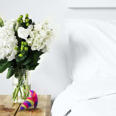 Colorful striped silicone plug resting on a wooden bedside table next to a vase of white flowers and a white bed.