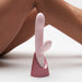 Blush pink dual-stimulation vibrator with ribbed shaft and clitoral arm, standing in a metallic pink charging base against a neutral background.
