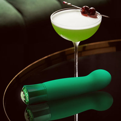 Green satin-smooth G-spot vibrator with faceted gem base next to a green cocktail on a reflective surface.