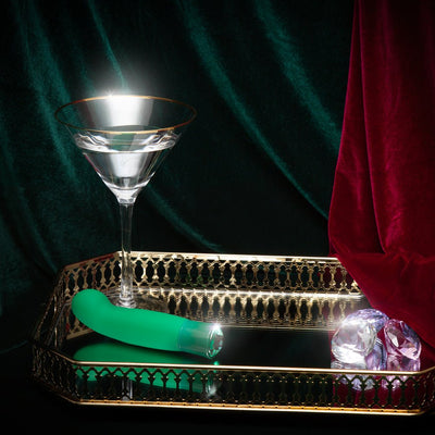Emerald green curved vibrator on gold tray with crystal accents and a martini glass against velvet backdrop