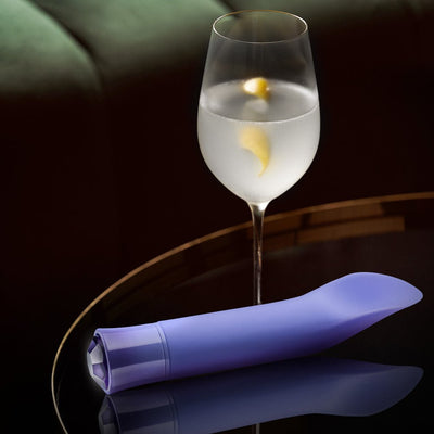 Sleek purple vibrator with faceted gem-like base resting on a reflective surface beside a glass of water with lemon peel.