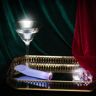 Lavender vibrator with faceted gem-like base on gold mirrored tray beside crystal accents and martini glass.