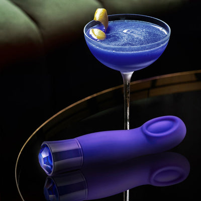 Purple sapphire-themed vibrator with faceted gem detail, displayed next to a blue cocktail on a reflective surface.