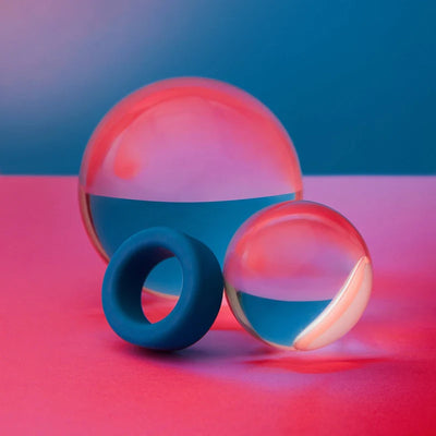 Blue silicone cockring displayed with two reflective glass spheres on a pink and blue background.