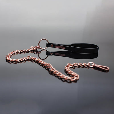 Rose gold chain leash with black vegan leather wrist strap and metal COQUETTE charm on reflective surface.