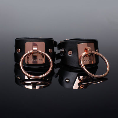 Pair of black vegan leather cuffs with rose gold metal rings and engraved Coquette plates on reflective black surface.