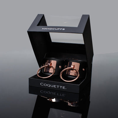 Coquette black vegan leather cuffs with rose gold metal rings in a black display box with clear lid and white lettering.
