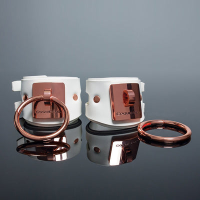 White vegan leather cuffs with rose gold metal rings and engraved Coquette plates on reflective surface