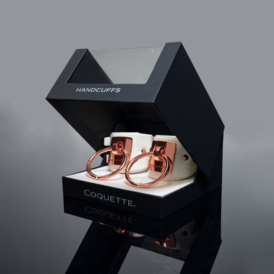 Coquette Pleasure Collection white vegan leather cuffs with rose gold metal rings in black display box labeled handcuffs.