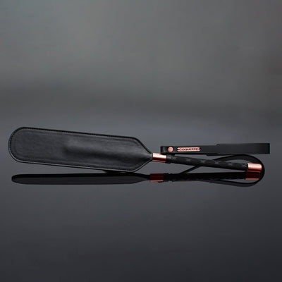 Black vegan leather paddle with diamond-patterned handle, rose gold accents, wrist strap, and Coquette metal charm on reflective surface.
