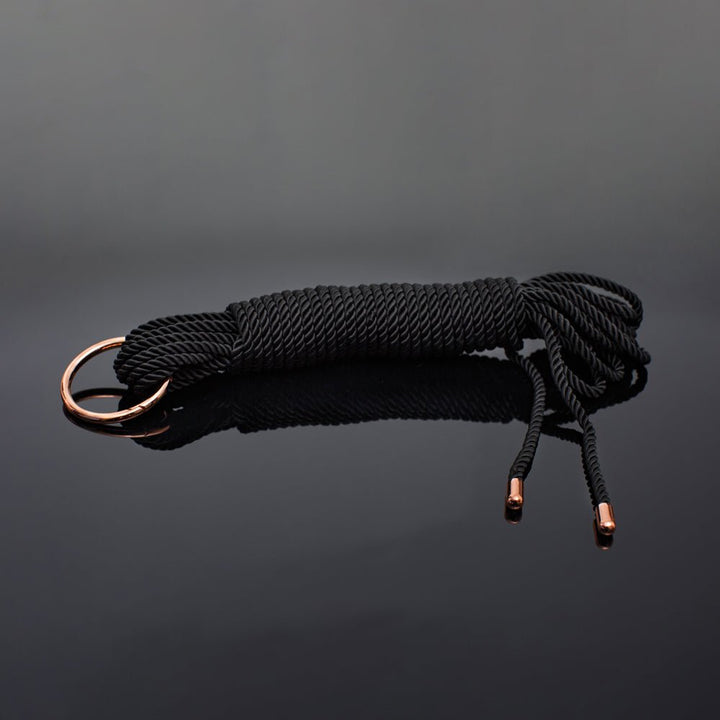 Coquette Pleasure Collection silky black rope with rose gold metal ring and tips on reflective black surface.