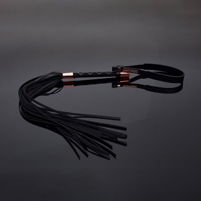 Black vegan leather whip with multiple tails, textured handle, rose gold accents, and wrist strap on reflective surface.