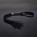 Black vegan leather whip with multiple tails, textured handle, rose gold accents, and wrist strap on reflective surface.