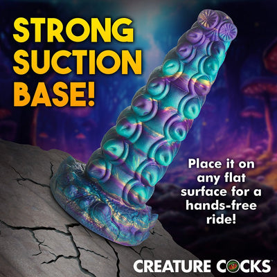 Iridescent teal, purple, and gold segmented fantasy adult toy with suction cup base on cracked stone surface.