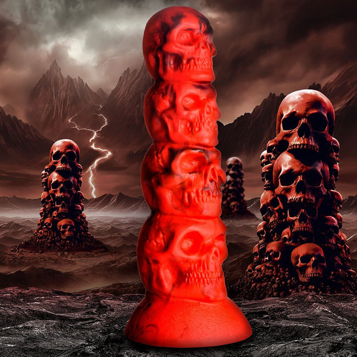 Red silicone dildo shaped as a vertical stack of four detailed skulls with a strong suction cup base against a dark, stormy mountain background.
