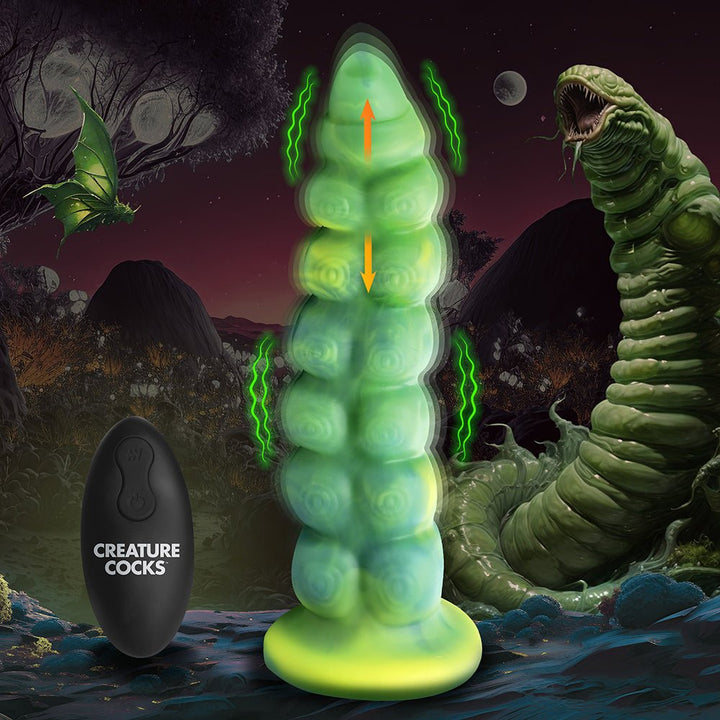 Green and yellow textured creature-inspired dildo with suction base and black remote control on a fantasy alien landscape background.