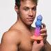 Shirtless man holding a colorful silicone fantasy dildo with blue, purple, and pink hues and a flared base.