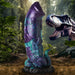 Dinosaur-themed adult toy in shimmering blue, purple, and teal with textured details and suction base, set against a jungle backdrop.