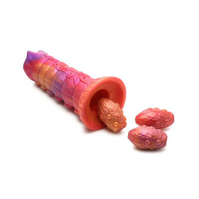 Sunset-colored silicone ovipositor dildo with textured segmented shaft and three soft, yellow-orange silicone eggs on white background.
