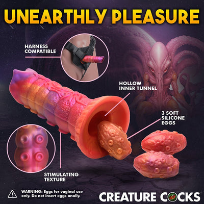 Orange and pink silicone ovipositor dildo with hollow tunnel, textured shaft, three soft eggs, and harness compatibility shown in sci-fi setting.