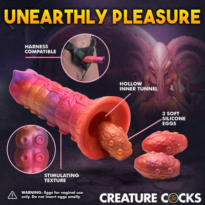 Orange and pink silicone ovipositor dildo with hollow tunnel, three soft eggs, textured surface, and harness compatibility details.